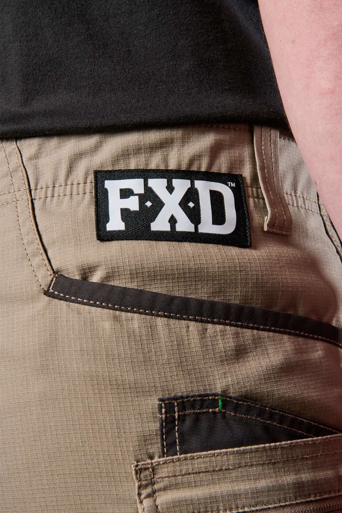 Close-up of an individual sporting the FXD WS-6 Work Short in beige, showcasing a black patch with "FXD" lettering on the back pocket. Renowned for their comfort and practicality, these shorts feature visible stitching and a loop located above the pocket.