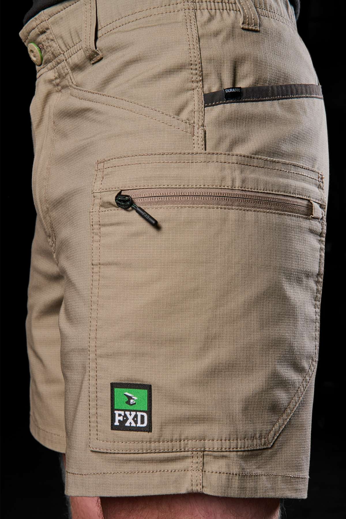 A close-up showcases an individual donning the khaki FXD WS-6 Work Short, celebrated for its comfort and functionality with several pockets. The side pocket is accented by a small green and black FXD logo, while the dark backdrop emphasizes the design and texture of these cargo shorts.