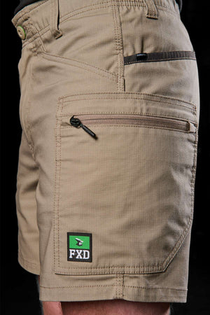 A close-up showcases an individual donning the khaki FXD WS-6 Work Short, celebrated for its comfort and functionality with several pockets. The side pocket is accented by a small green and black FXD logo, while the dark backdrop emphasizes the design and texture of these cargo shorts.