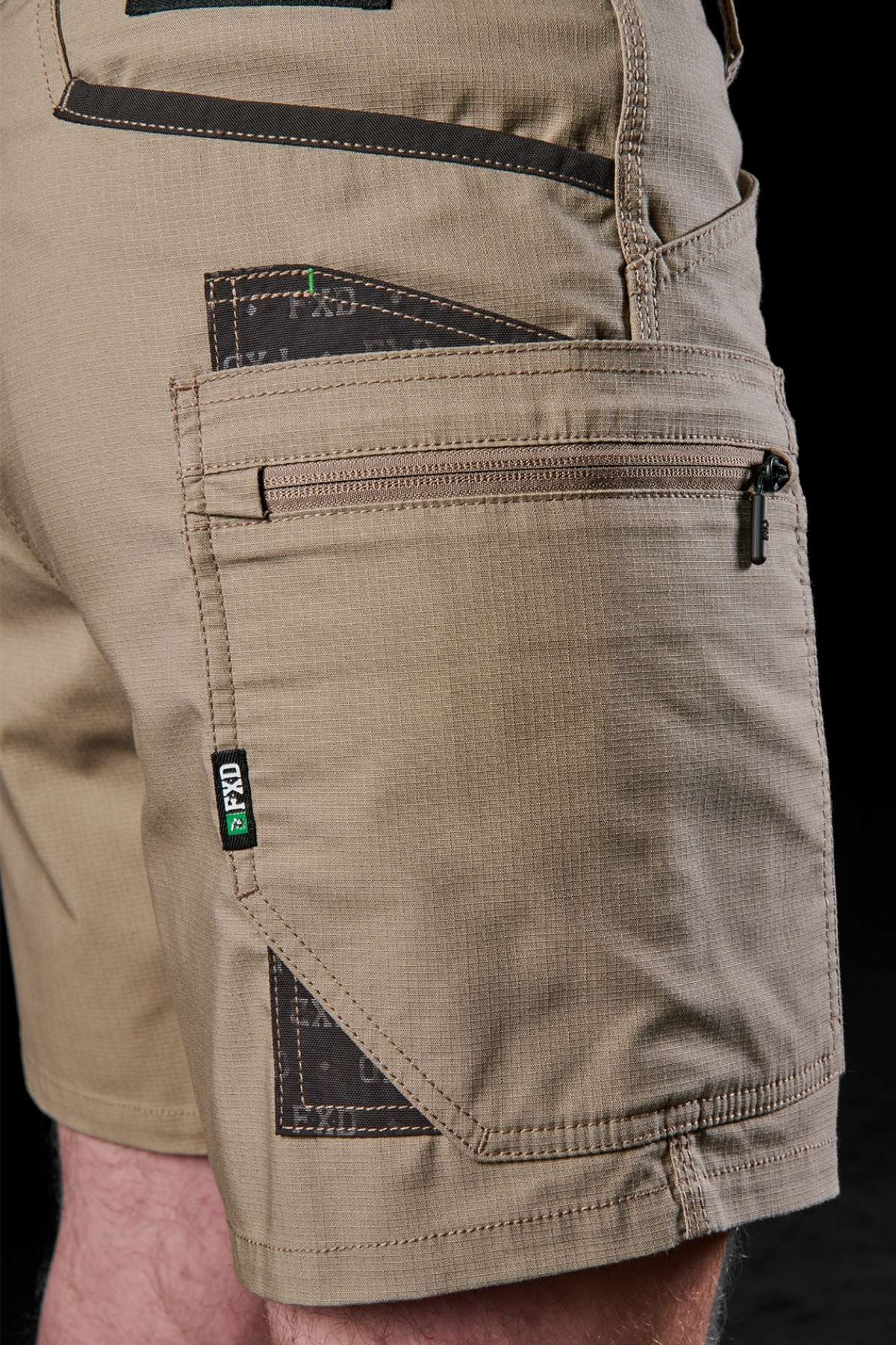 Close-up of a person wearing FXD WS-6 Work Short in khaki, designed with several pockets including a secure zippered pocket. The shorts offer comfort and practicality, highlighted by dark patches and a logo tag. The black background accentuates the intricate texture and fabric design.