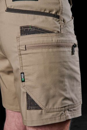 Close-up of a person wearing FXD WS-6 Work Short in khaki, designed with several pockets including a secure zippered pocket. The shorts offer comfort and practicality, highlighted by dark patches and a logo tag. The black background accentuates the intricate texture and fabric design.