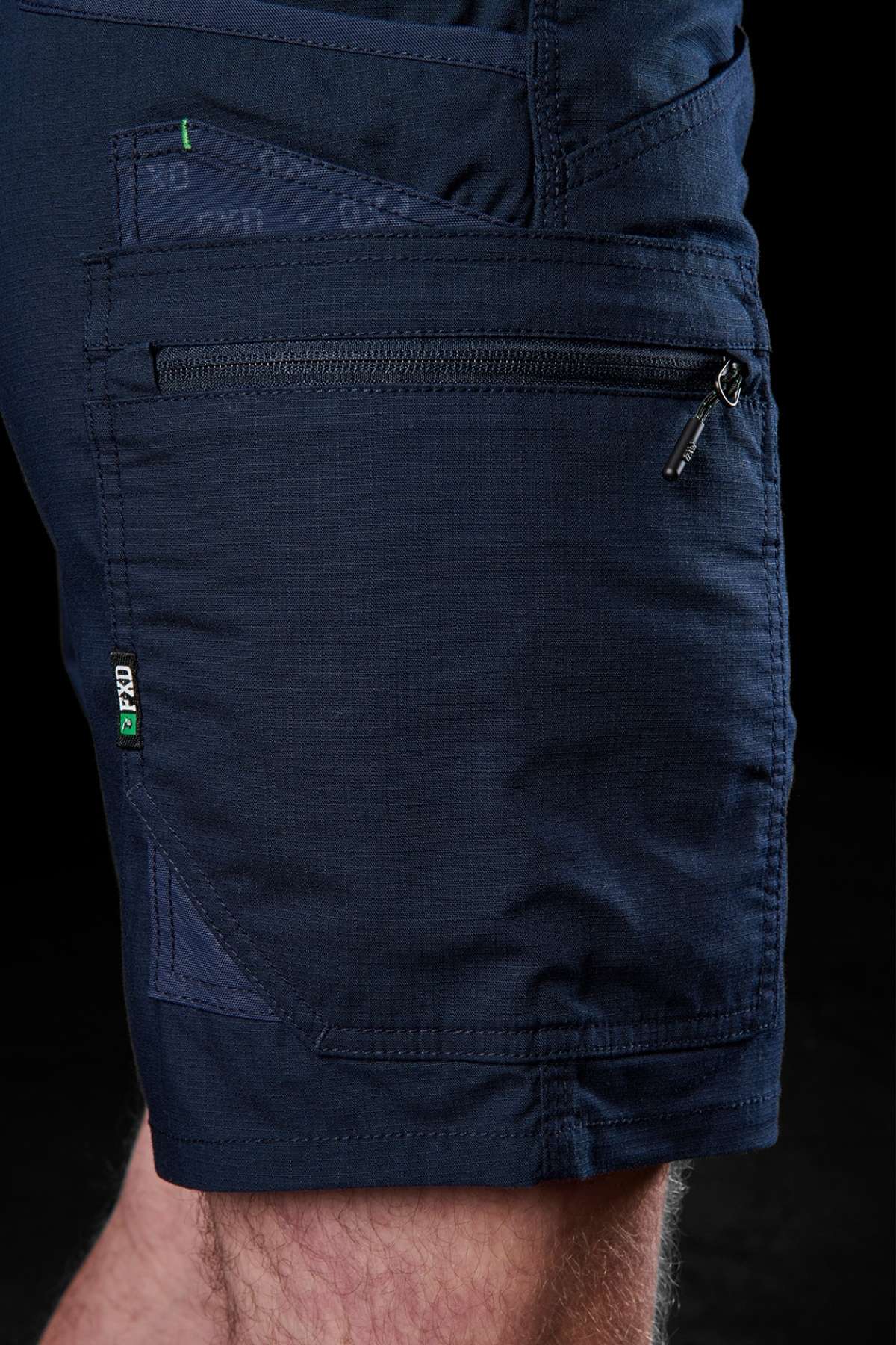 A close-up shot shows someone wearing the FXD WS-6 Work Short in dark navy, featuring a zippered pocket. The textured fabric and visible stitching emphasize the added stretch, while a small green and white logo tag on the seam seamlessly merges utility with style.