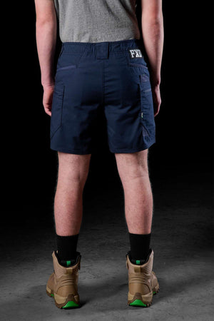 A person stands facing away, wearing a gray shirt and navy blue FXD WS-6 Work Shorts with added stretch and the letters "FDL" on the back. Tan boots with green soles add contrast to black socks, all set against a dark background.