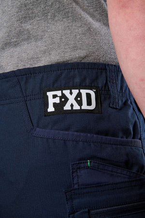 A close-up shows someone in a grey shirt and FXD WS-6 Work Shorts in dark blue, which offer added stretch for comfort and display a black and white "FXD" label near the pocket.