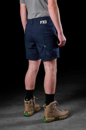 A person in a gray shirt, dark blue FXD WS-6 Work Short showcasing "FXD" on the back, black socks, and tan boots stands against a shadowy backdrop with their back to the camera. The stretch feature of the shorts suggests enhanced comfort and durability.