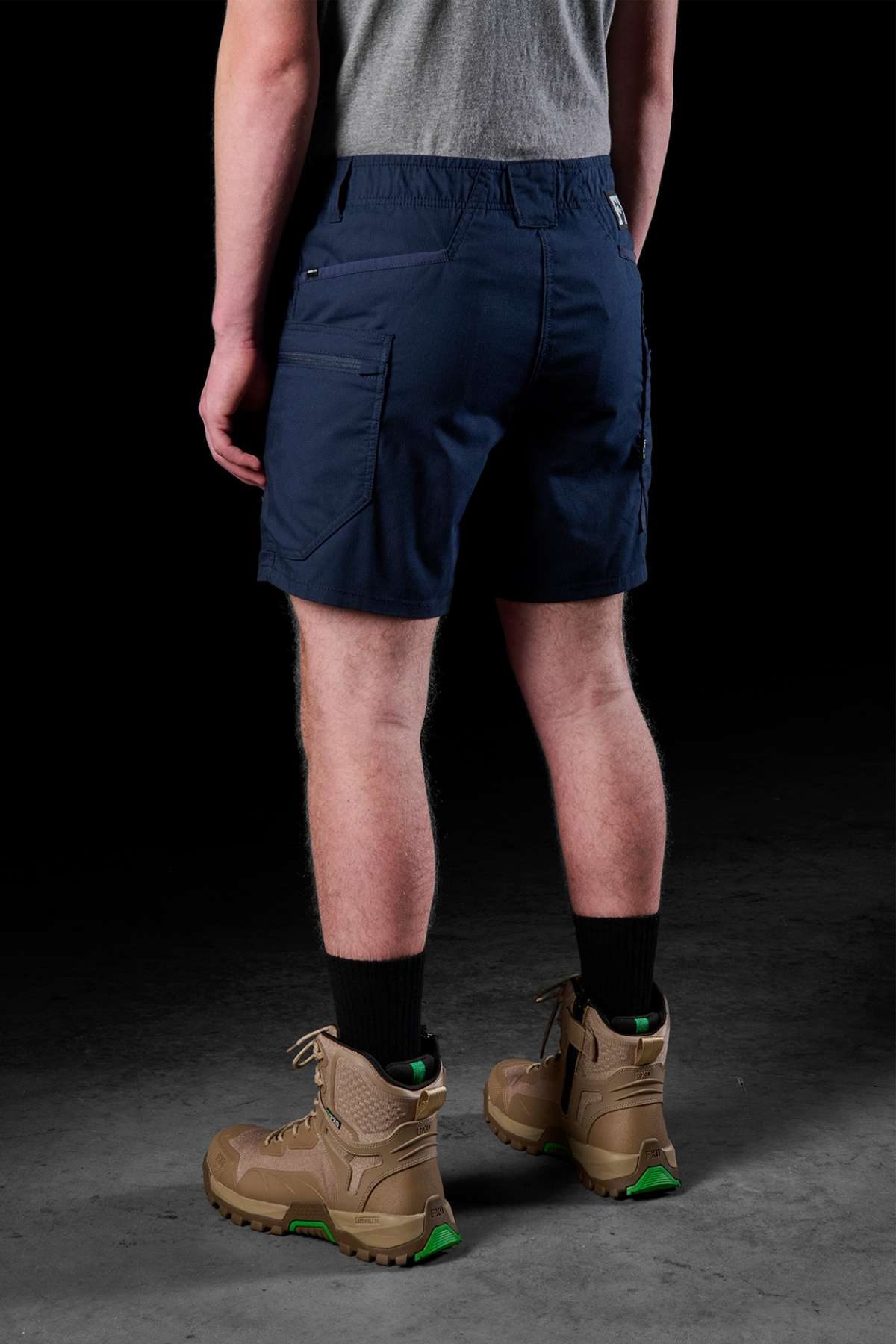A person faces away, showcasing a gray shirt and the FXD WS-6 Work Short, designed with multiple pockets for enhanced flexibility. Black socks are visible above tan hiking boots with green soles, contrasted against a dark background.