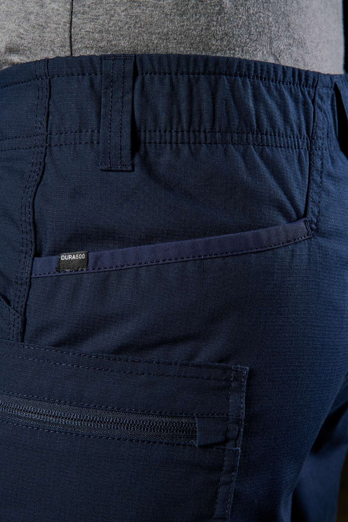 Close-up of a person showcasing the FXD WS-6 Work Short in navy blue, featuring pocket and stitching details. The fabric tag displays "DURA500." These cargo shorts offer added stretch, a textured finish, and include a visible zipper pocket. The individual is paired with a gray shirt.
