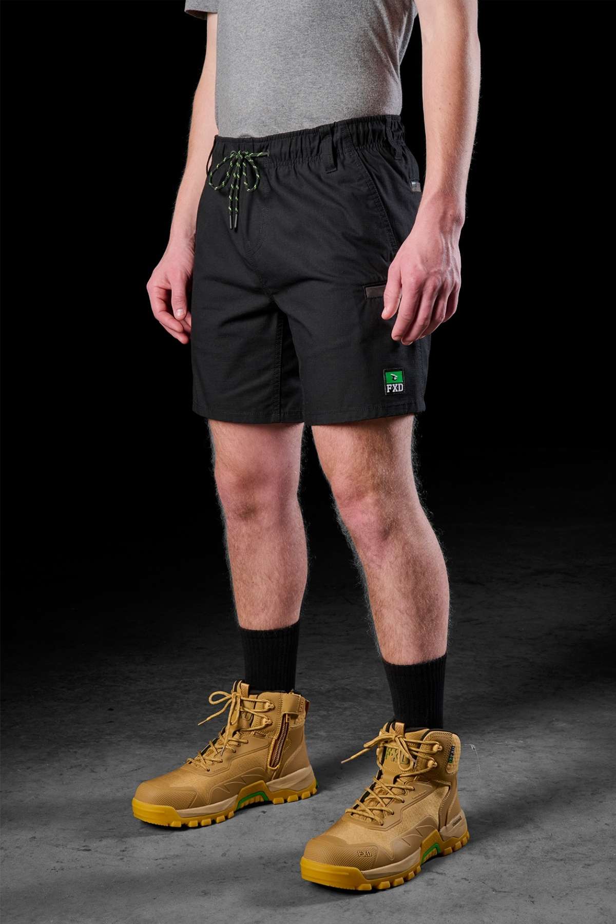 A person is wearing a gray shirt and the FXD WS-7 Work Shorts against a dark background. These tough shorts boast an elastic waistband and display a discreet green and black logo on the left leg, complemented by beige work boots and black socks.