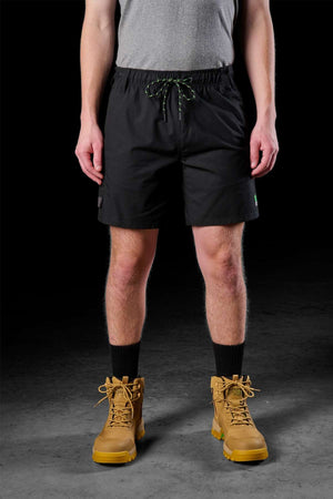 A person wearing a gray shirt and the FXD WS-7 Work Short in black, featuring an elastic waist for durability, stands against a dark background. Tan lace-up boots paired with black socks just above complete the look as their hands rest casually at their sides.