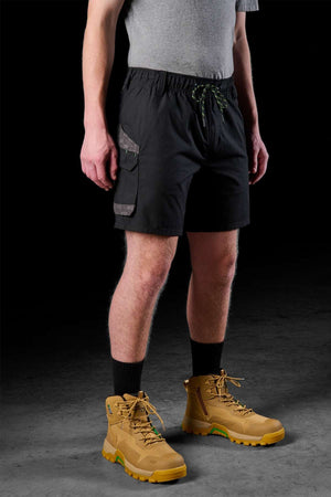 A person dressed in a gray t-shirt, FXD WS-7 Work Short with side pockets, tall black socks, and tan hiking boots accented with green stands against a dark background. These sturdy shorts include an elastic waistband ideal for both comfort and style.