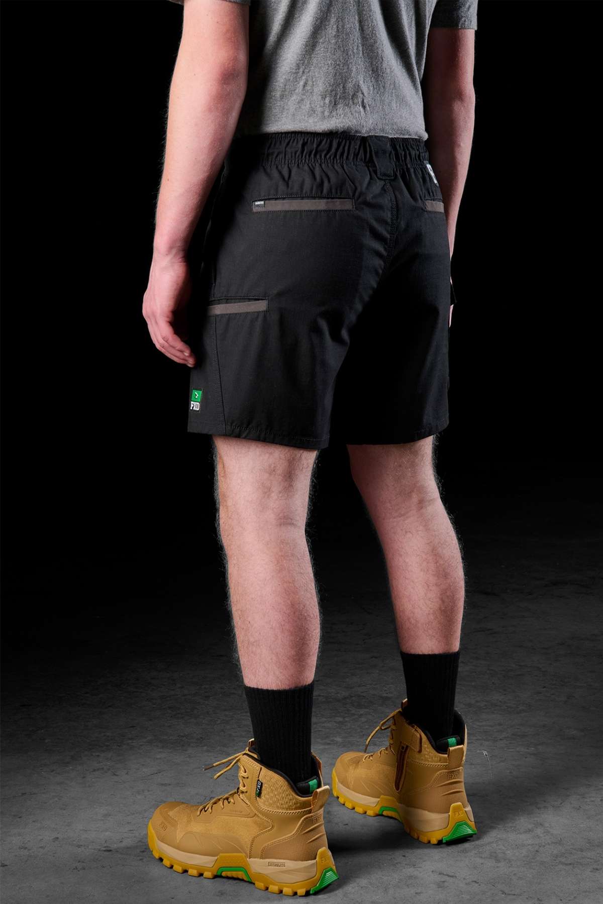 A person wearing a gray shirt, FXD WS-7 Work Short in black, and tan work boots with green soles stands on a dark surface. Their back is facing the camera, and the lighting highlights the texture of the clothing and boots.