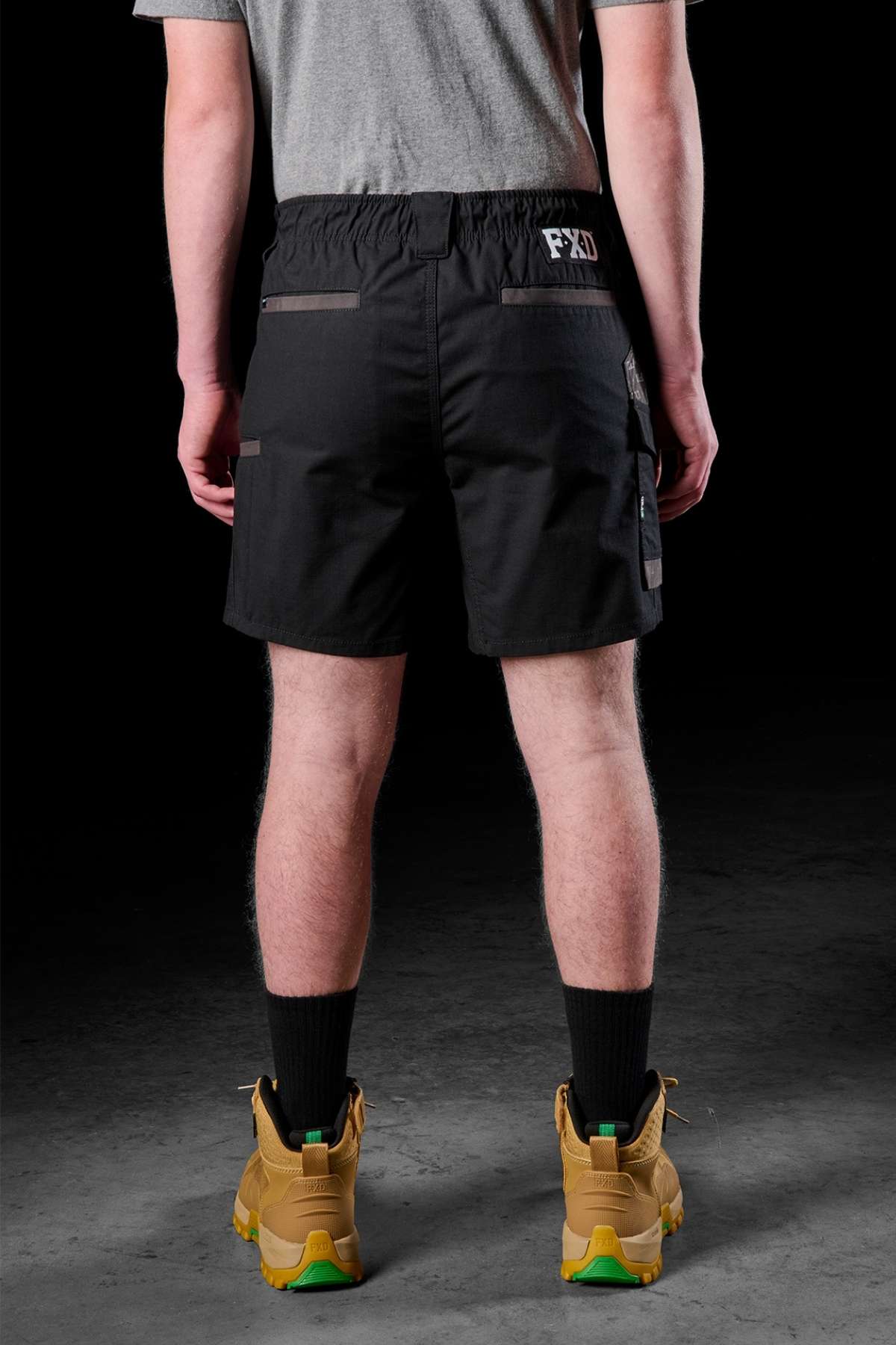 Rear view of a person wearing a gray shirt and the durable FXD WS-7 Work Short featuring an elastic waist, along with black socks and tan work boots with green soles, standing on a dark gray surface.