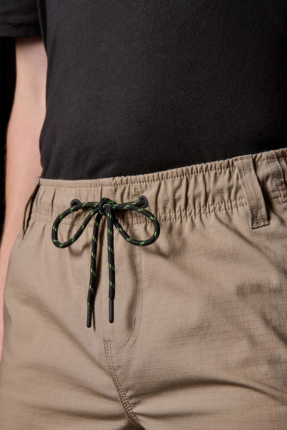 Close-up of a person wearing a black shirt and beige FXD WS-7 Work Short, highlighting the comfort and utility of these versatile bottoms. The shorts feature an elastic waistband with a black and green drawstring tied in a bow, one hand casually slipped into a pocket, all while sporting a subtle grid pattern.
