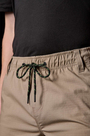 Close-up of a person wearing a black shirt and beige FXD WS-7 Work Short, highlighting the comfort and utility of these versatile bottoms. The shorts feature an elastic waistband with a black and green drawstring tied in a bow, one hand casually slipped into a pocket, all while sporting a subtle grid pattern.