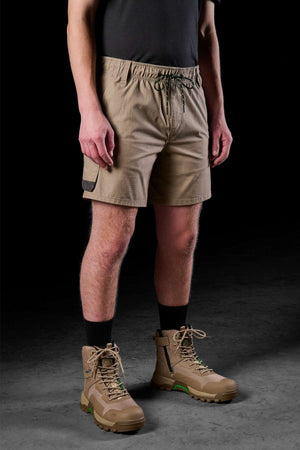A person sporting FXD WS-7 Work Shorts in beige, paired with a dark shirt, black socks, and tan hiking boots is set against a dark backdrop. This ensemble highlights a casual outdoor style while providing enhanced comfort and utility for any adventure.