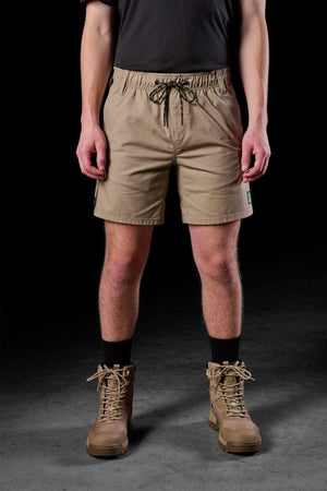 A person is dressed in a black t-shirt matched with the FXD WS-7 Work Short for maximum comfort, along with black socks and tan hiking boots, set against a dark background.