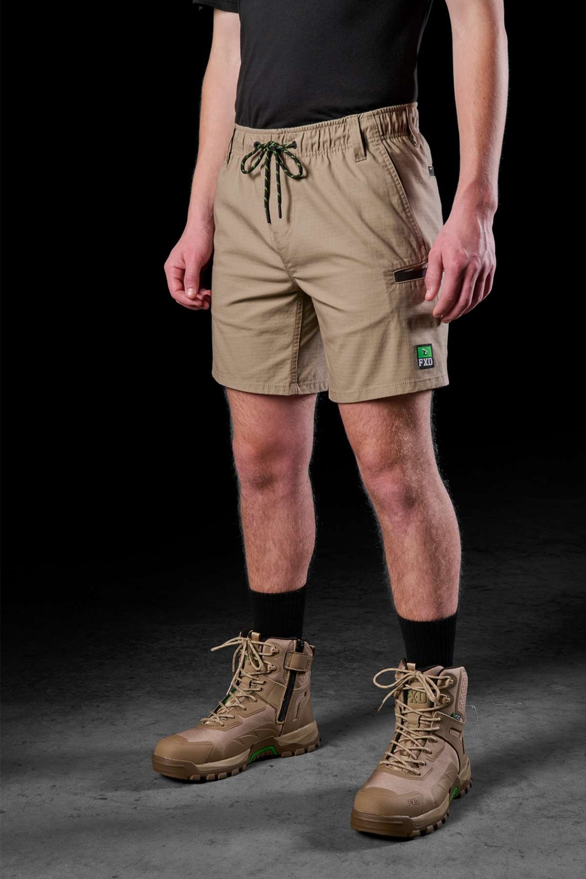 In a dim environment, an individual radiates a sense of ease and practicality. They're dressed in a black T-shirt, FXD WS-7 Work Shorts in tan featuring a drawstring, complemented by black socks and tan hiking boots. The shorts have a small text patch on the pocket, while the person faces the camera with arms at ease.