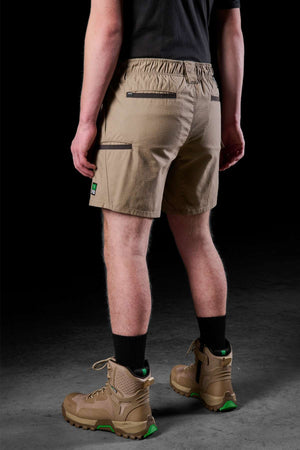 A person dressed in a black shirt and FXD WS-7 Work Short, known for its utility and comfort, is matched with black socks and tan hiking boots with green soles, standing against a dark background. The emphasis is on the back view of the shorts and boots.
