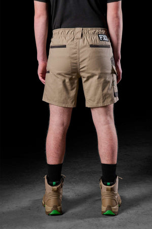 A person, prioritizing comfort and practicality, dons a black shirt paired with the FXD WS-7 Work Short in tan, which boasts numerous pockets and a back patch. Black socks are visible as they rise from robust tan boots while the individual stands with their back to the camera against a dim backdrop.