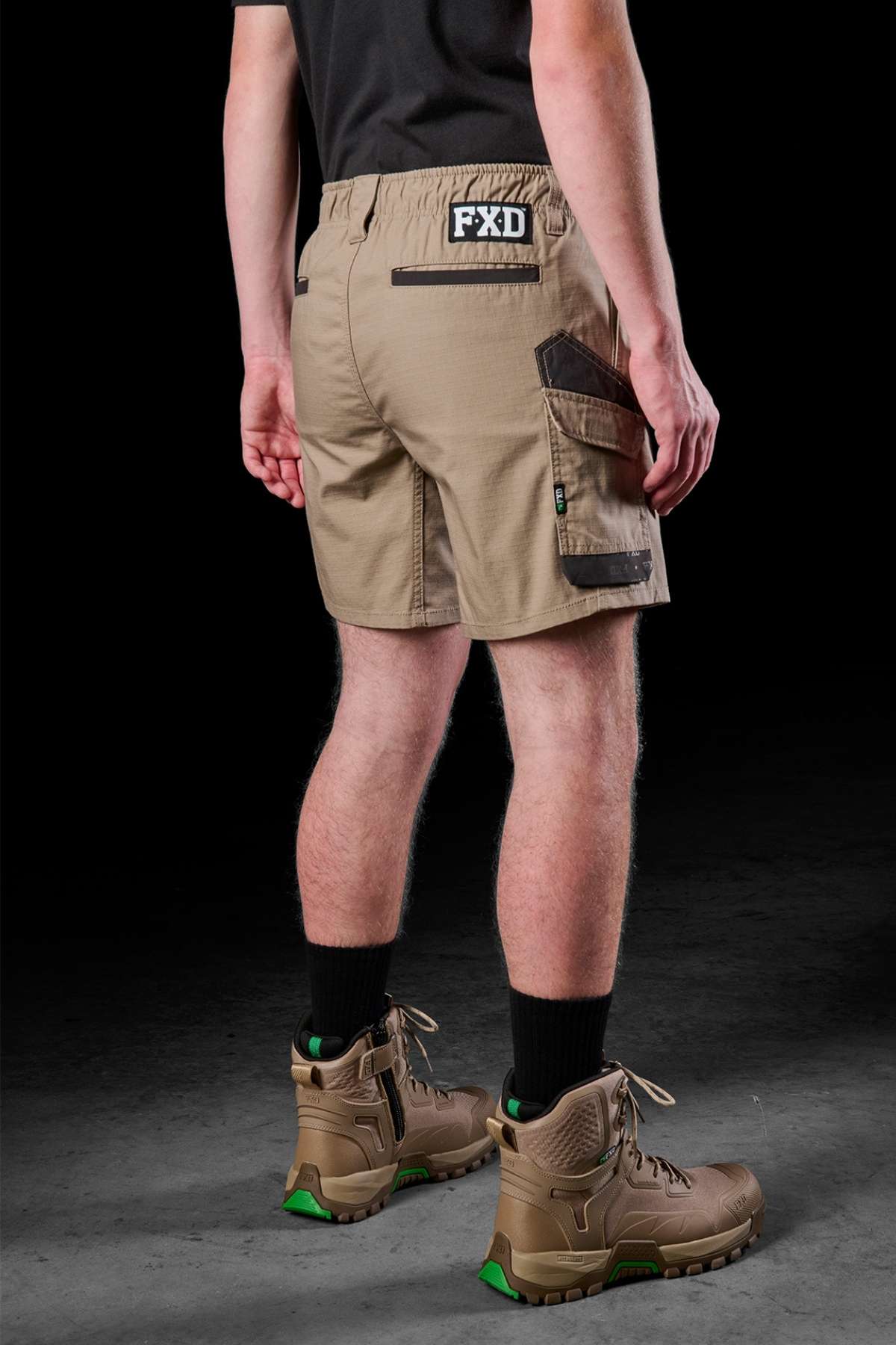 With their back turned, a person is wearing tan FXD WS-7 Work Shorts, which display "FXD" on the waistband and pockets accented with black trim. These shorts combine utility with comfort. They complete their outfit with black socks and brown high-top boots featuring green accents, all set against a dark background.