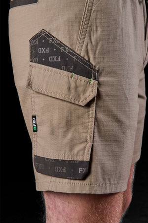 A close-up shot captures a person wearing the FXD WS-7 Work Short in beige, emphasizing its comfort and practicality. The shorts showcase a black and gray pocket design adorned with the "FXD" logo, along with reinforced stitching and a flap-covered side pocket, all against a stylish black backdrop.