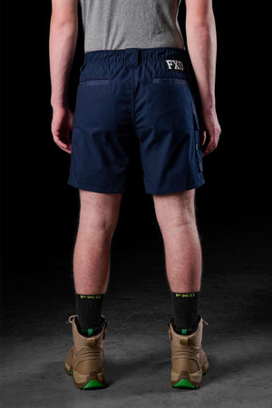 A person faces away from the camera, showcasing a gray shirt, FXD WS-7 work shorts in navy blue with the "FXD" label on the waistband, black socks, and tan work boots. This sturdy outfit projects comfort against the dark backdrop.