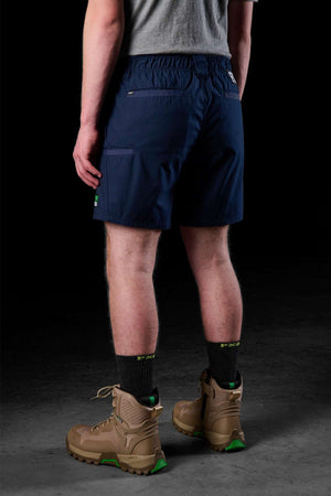 A person wearing a grey T-shirt, FXD WS-7 durable navy blue work shorts with pockets, black socks, and brown hiking boots with green accents stands against a dark background, facing away from the camera.