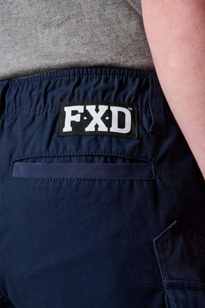 Close-up image of a person wearing the FXD WS-7 Work Short in navy blue, showcasing the "FXD" brand logo on the waistband. The shorts feature a back pocket with visible stitching, designed for comfort. The person is wearing a gray shirt tucked in.