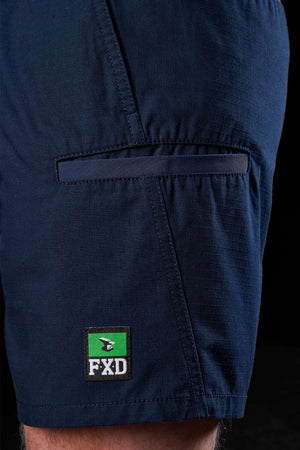 A close-up shot features someone wearing the FXD WS-7 Work Short, showcasing its durable dark blue fabric and a green and black FXD logo patch near the hem. The shorts highlight visible stitching and include a side pocket with a flap for comfort, set against a black background.