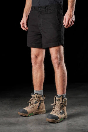 A person standing against a dark background, wearing FXD WS-2 Short Short, gray socks, and rugged tan work boots with black accents. Their hands are relaxed at their sides, and only the lower half of their body is visible.