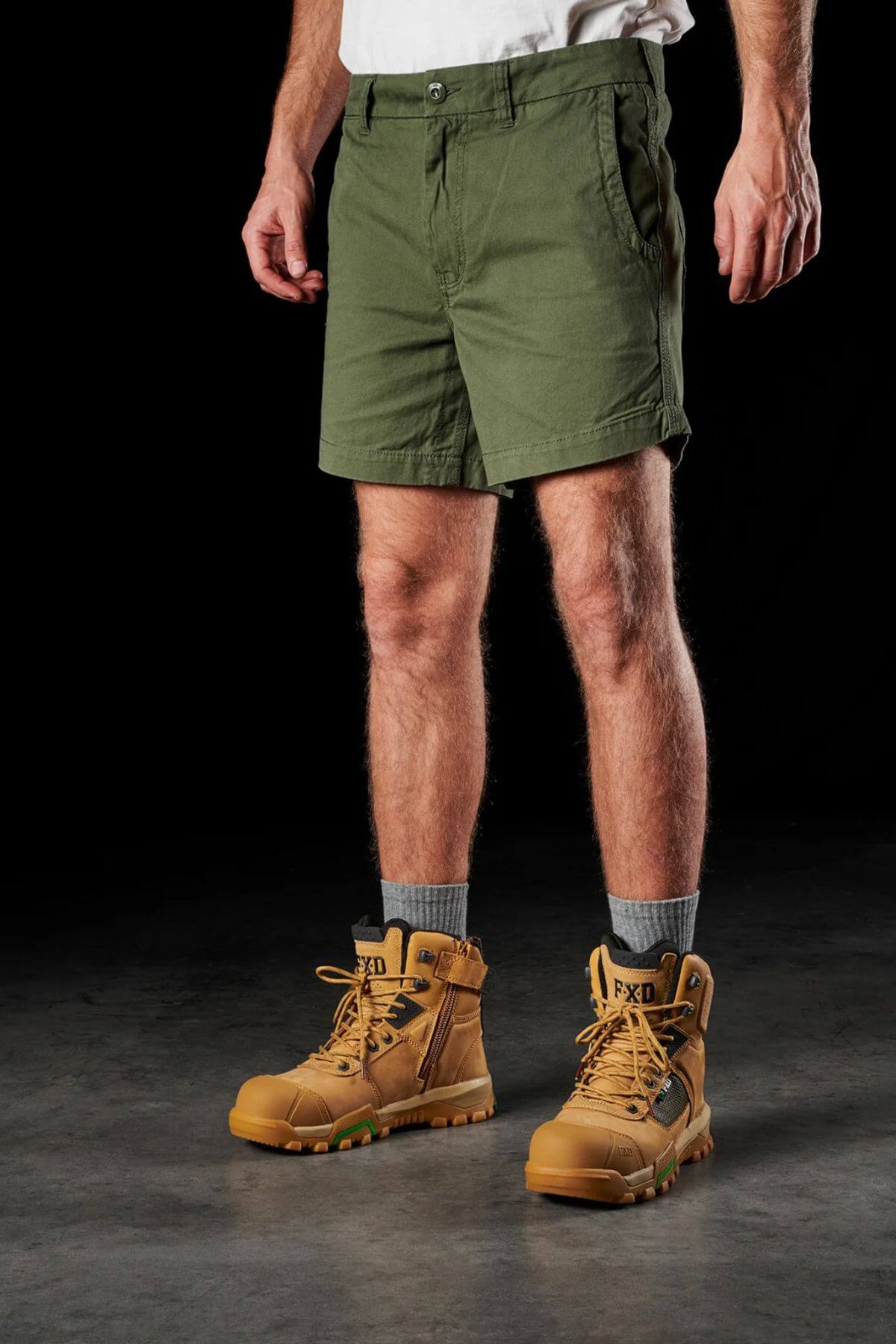 A person wearing a white T-shirt, FXD WS-2 Short Short, gray socks, and tan work boots stands against a dark background. Only the lower half of their body is visible. The boots have "FXD" labeled on the tongue and feature green accents on the soles.
