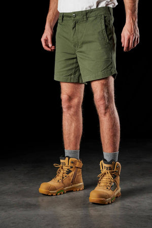 A person wearing a white T-shirt, FXD WS-2 Short Short, gray socks, and tan work boots stands against a dark background. Only the lower half of their body is visible. The boots have "FXD" labeled on the tongue and feature green accents on the soles.