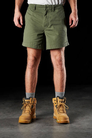 A person is shown from the waist down wearing a pair of green FXD WS-2 Short Shorts crafted from soft touch cotton twill, a white shirt tucked in, and tan work boots with gray socks. The black background highlights the casual and practical nature of the clothing attire.