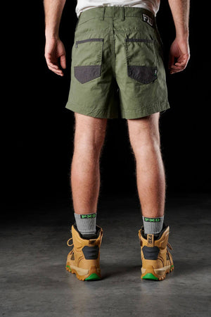 Rear view of a person wearing green FXD WS-2 Short Shorts, which have two pockets and an FXD logo, paired with gray socks and tan work boots featuring green soles. The shorts are made from soft touch cotton twill and include DURA500™ abrasion panels. The background is black.