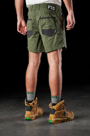 A person is seen from behind, wearing the FXD WS-2 Short Short in olive-green, made from soft touch cotton twill and featuring black pockets with DURA500™ abrasion panels. They are also wearing gray socks and brown work boots with black accents. The background is black, and the person stands on a gray concrete surface.