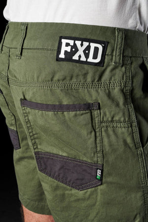 Close-up of the back pocket area of FXD WS-2 Short Shorts in green with black accents. The shorts feature a black patch on the waistband displaying the white letters "FXD," DURA500™ abrasion panels, and a smaller green and white label on the pocket. The bottom part of a white shirt is also visible.