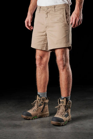 A person stands against a dark background, wearing FXD WS-2 Short Shorts made from soft-touch cotton twill, paired with gray socks and sturdy brown work boots featuring black and green accents. Their upper body is not visible. The floor beneath them is smooth and gray.