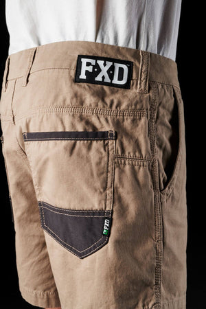 A close-up of a person wearing FXD WS-2 Short Shorts crafted from soft-touch cotton twill, showcasing multiple pockets with black detailing. The back pocket is highlighted by a black accent and prominently displays a patch with "FXD" above it. The person pairs the shorts with a white shirt tucked in.