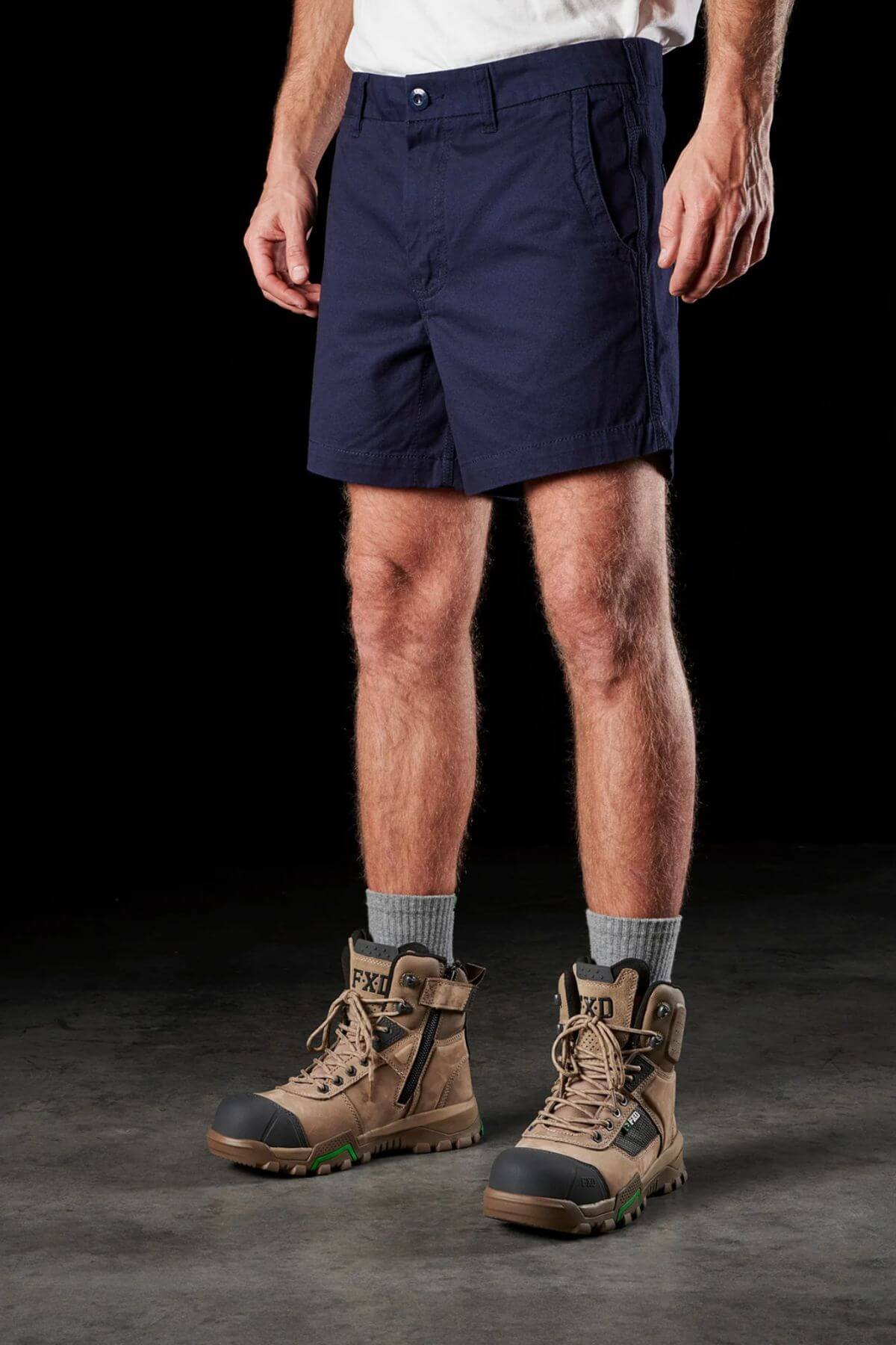 A person stands against a black background, showcasing the lower half of their body. They wear FXD WS-2 Short Shorts in navy blue, light gray socks, and brown work boots.