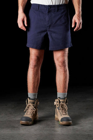 A person wearing FXD WS-2 Short Shorts in durable dark blue, a white shirt, gray socks, and tan work boots stands against a black background. Only the lower part of the body is visible in the image.