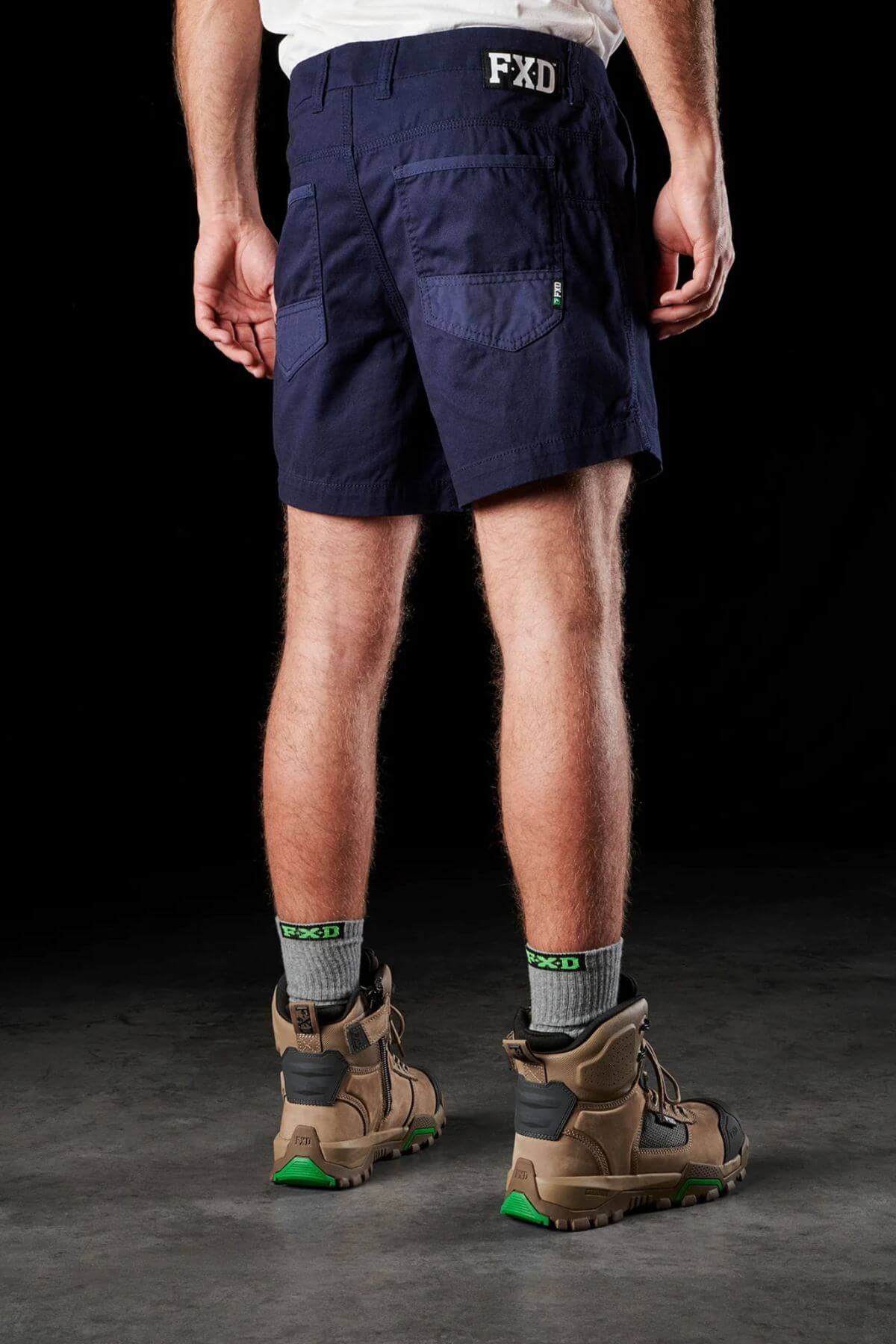 A person stands with their back to the camera, wearing the FXD WS-2 Short Short in navy blue, featuring "FXD" initials on the waistband. They complement the shorts with gray socks adorned with "FXD" logos and brown work boots accented in green. The black background accentuates their stylish attire and footwear.