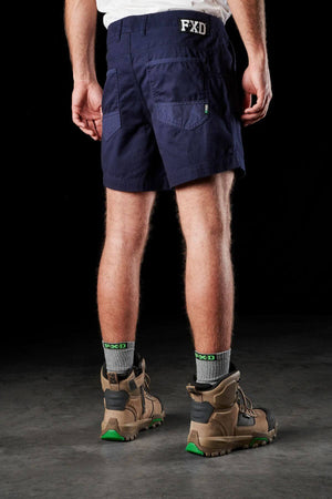 A person stands with their back to the camera, wearing the FXD WS-2 Short Short in navy blue, featuring "FXD" initials on the waistband. They complement the shorts with gray socks adorned with "FXD" logos and brown work boots accented in green. The black background accentuates their stylish attire and footwear.