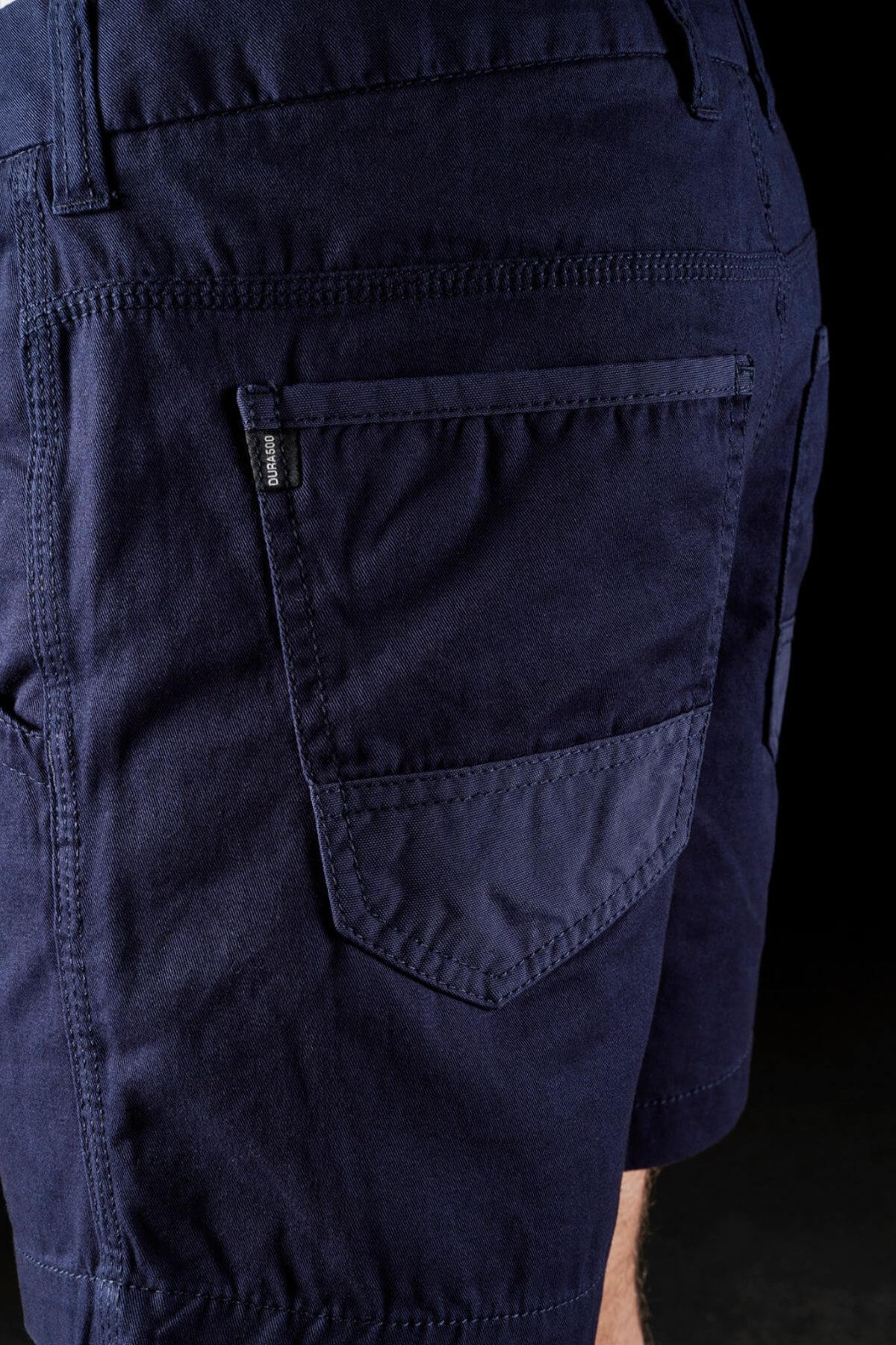 A close-up view showcases a person wearing the FXD WS-2 Short Short in dark blue. The image focuses on the back pocket, highlighting detailed stitching and a small black tag near the pocket's seam. The black background accentuates the fabric's color and texture.