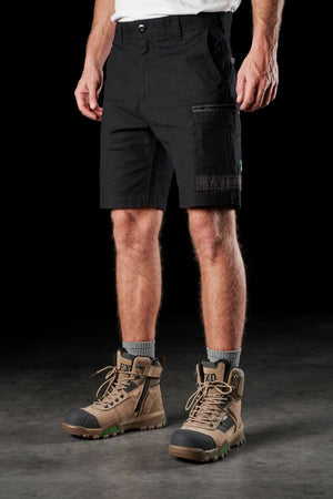 A person wearing FXD WS-3 Shorts made from stretch cotton canvas stands against a dark background. They have on light brown hiking boots with green accents and gray socks pulled up to the ankles. The durable shorts feature Dura500™ fabric and triple needle seams, ensuring both comfort and resilience.