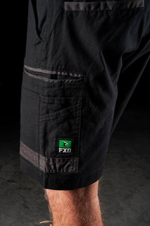Close-up view of a person wearing FXD WS-3 Shorts, crafted from stretch cotton canvas with reinforced triple needle seams and equipped with multiple pockets. A small green label displaying the text "FXD" adorns the side pocket. The background is dark and indistinct.