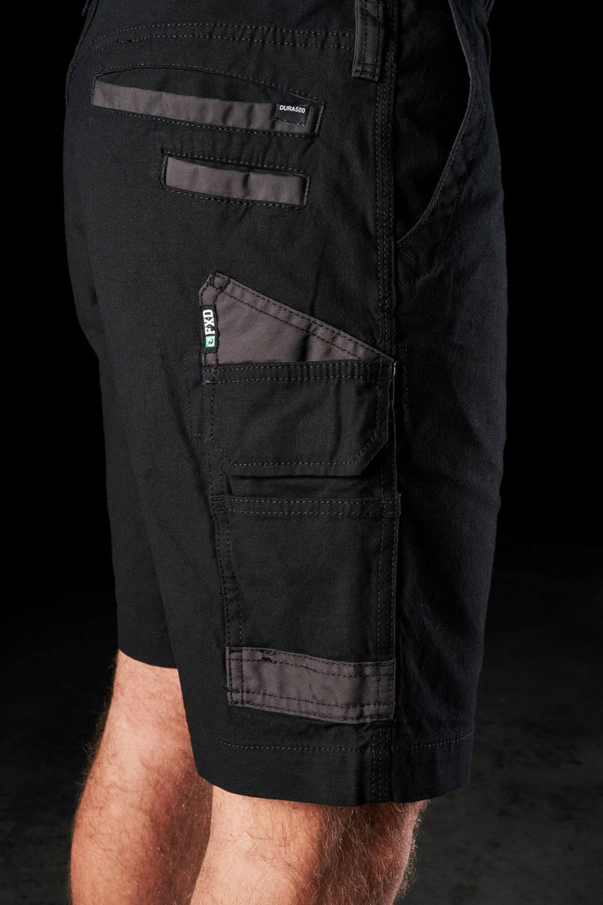 A close-up of a person wearing the FXD WS-3 Shorts made from black stretch cotton canvas, showcasing multiple zippered and velcro pockets. The image focuses on the thigh and hip area, emphasizing the practical design with triple needle seams and detailed stitching against a dark background.