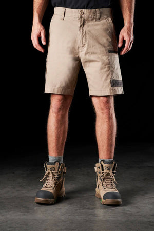 A person is wearing beige FXD WS-3 Shorts made from stretch cotton canvas, grey socks, and tan work boots featuring black and green details. They are standing against a plain black background with no other items or accessories visible.