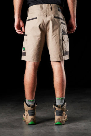 Rear view of a person wearing stretch cotton canvas FXD WS-3 Shorts in beige, along with hiking boots and black-green socks. The background is black, and the person's arms are relaxed by their sides.