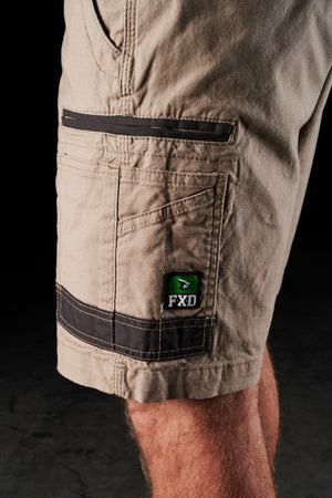 Close-up of a person’s leg wearing FXD WS-3 Shorts crafted from stretch cotton canvas in beige, showcasing multiple pockets. The shorts feature a small green and black patch with "FXD" on the pocket and are reinforced with triple-needle seams. A dark background accentuates the details of the clothing.