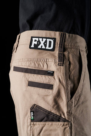 Close-up of a person wearing FXD WS-3 Shorts with brown stitching and multiple pockets. The shorts, crafted from stretch cotton canvas, feature a tag with the logo "FXD" on the waistband and another smaller tag that reads “DURA500” on a pocket. Triple needle seams add durability. The background is black.